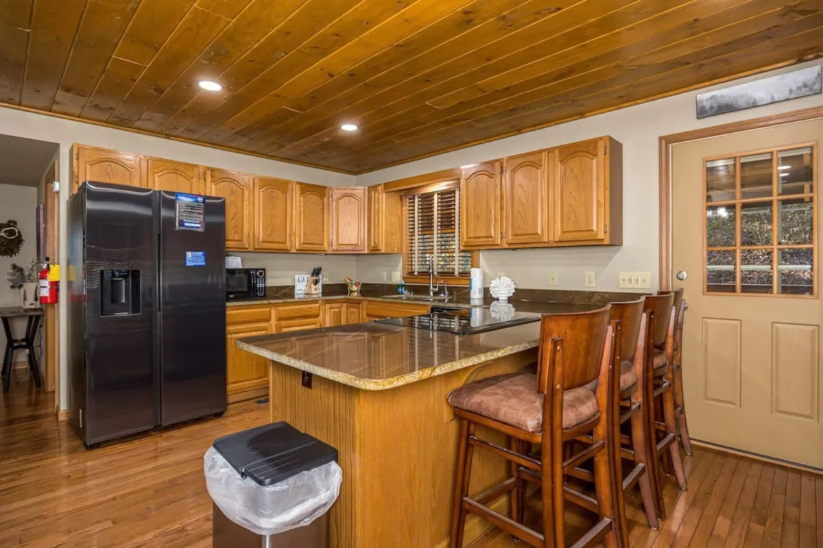 A bright kitchen area with a central island, bar stools, wooden cabinets, and a window letting in plenty of natural light. The setting encourages casual conversations, shared cooking, and meals with friends and family.