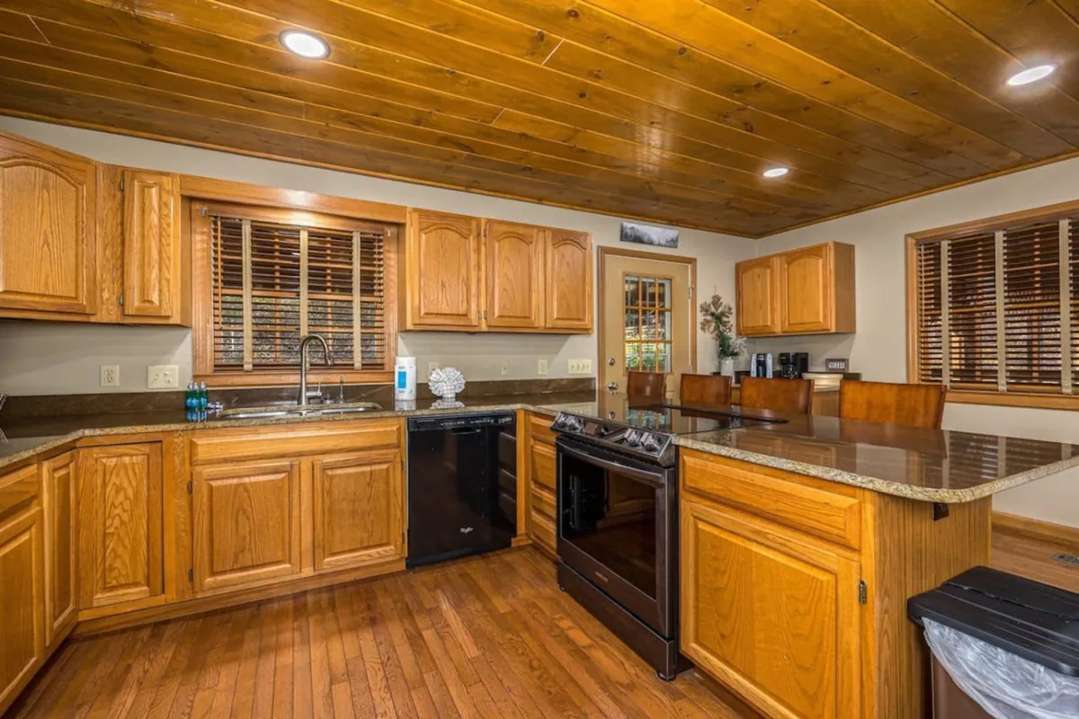 This image showcases a warm, inviting kitchen with wooden cabinets, granite countertops, and stainless steel appliances. The layout is spacious, allowing for multiple people to move and cook comfortably. The modern appliances and ample counter space are complemented by cozy, homey touches that suggest gatherings and shared meals.