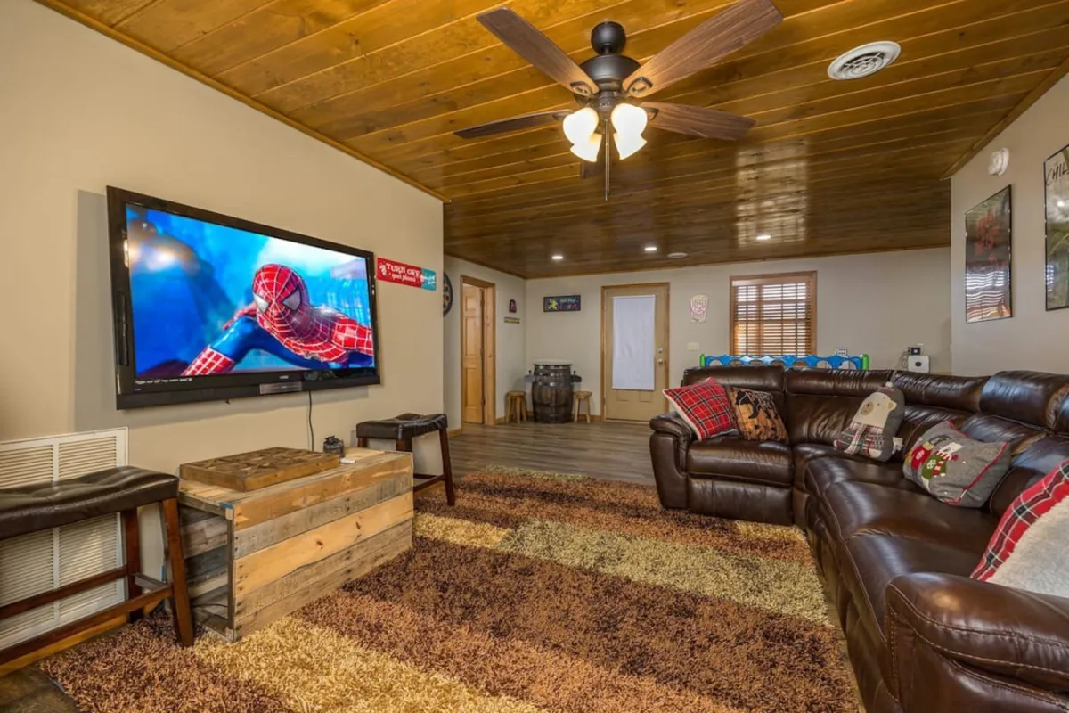 A spacious and cozy living area indoors with a large television displaying a Spider-Man scene, comfortable leather sofa, and rustic wooden coffee table. The mood is relaxed and playful, perfect for family gatherings or game nights after a day exploring Gatlinburg.