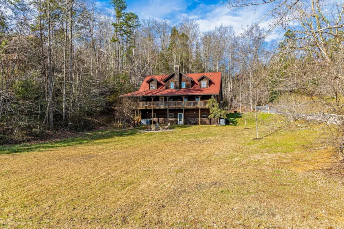 This exterior image showcases a welcoming log cabin structure with a distinctive red roof, set amid a spacious front lawn and surrounded by trees. The setting is open and accessible, allowing for easy parking and outdoor gatherings.