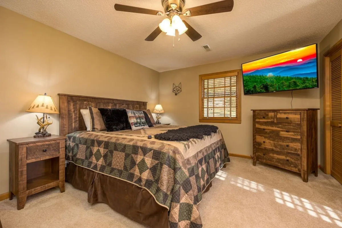 A comfortable bedroom highlighted by a bed with a quilt, a mounted TV, a nightstand with a lamp, and a dresser. The room is designed for relaxation and quiet moments, perfect for both winding down in the evening and lazy mornings.