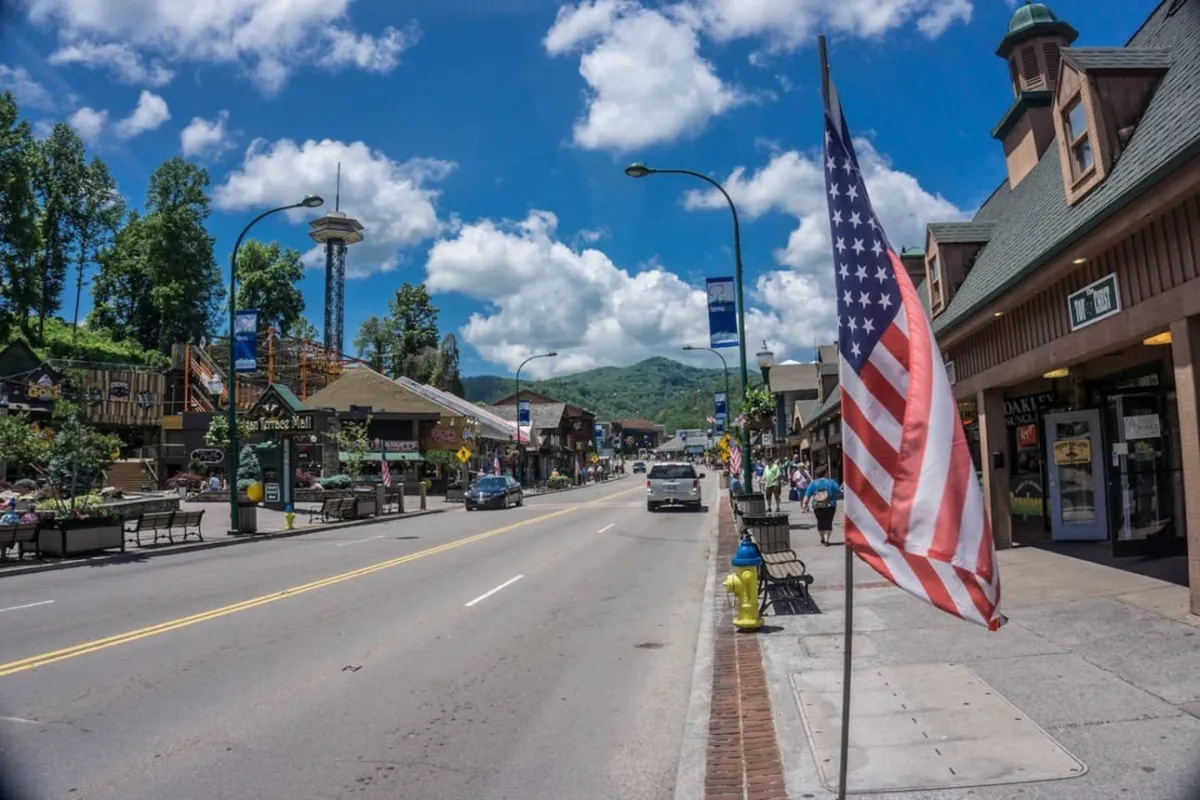 A lively street scene featuring an American flag, quaint shops, and a backdrop of the Smoky Mountains, bustling with people shopping, dining, and sightseeing. The festive atmosphere and town square reflect a vibrant downtown experience.