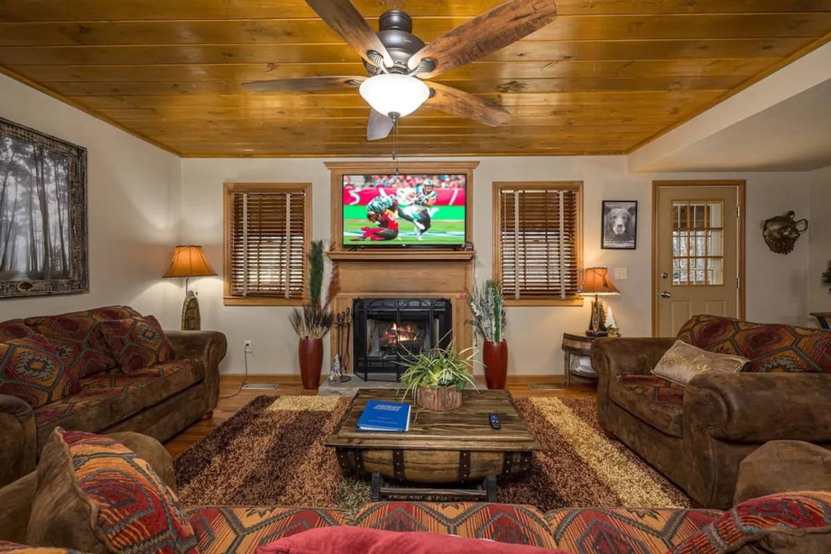 A comfortable living space with sofas arranged around a coffee table, a fireplace, a television, and warm wooden ceilings. The setup is ideal for group movie nights, game sessions, or relaxing chats by the fire, with expansive windows that let in plenty of daylight.