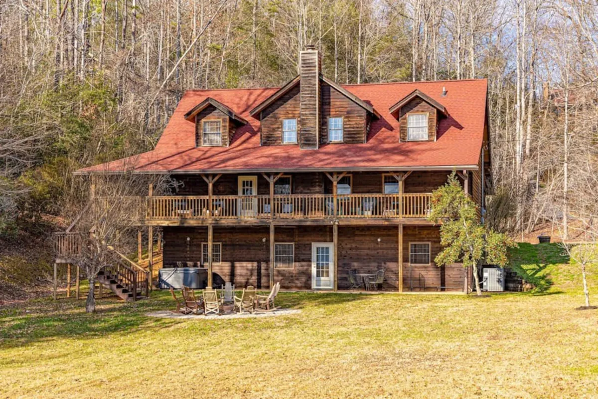 A warm and welcoming cabin exterior with a red roof, spacious front porch, and a fire pit area set against a backdrop of trees. The scene suggests space for outdoor gatherings, family storytelling, and enjoying evenings together under the stars.