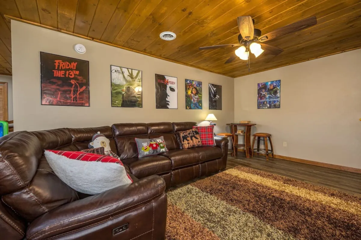 A comfortable media nook with a leather sofa, movie posters, ceiling fan, and coffee table, inviting guests to lounge and enjoy movie nights or relax between games. The mood is laid-back, with clear references to both movie and gaming activities.