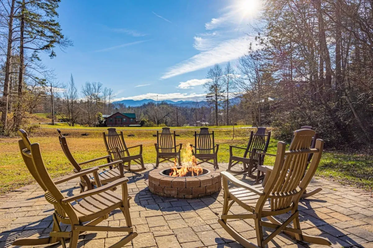 The image showcases an inviting outdoor fire pit surrounded by classic wooden rocking chairs, set against a breathtaking backdrop of the Smoky Mountains. The setting suggests cozy evenings spent sharing stories, roasting s’mores, and enjoying the sweeping mountain views as the sun sets.