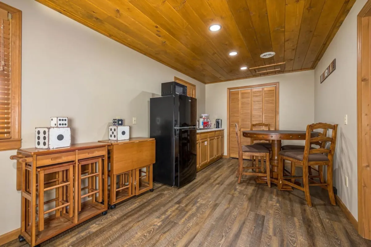 The image features a casual dining nook within the kitchen, with black refrigerator, wooden bar stools at a counter, and a small table in the background. The setup invites casual gatherings and shared snacks, with a warm and inviting atmosphere.