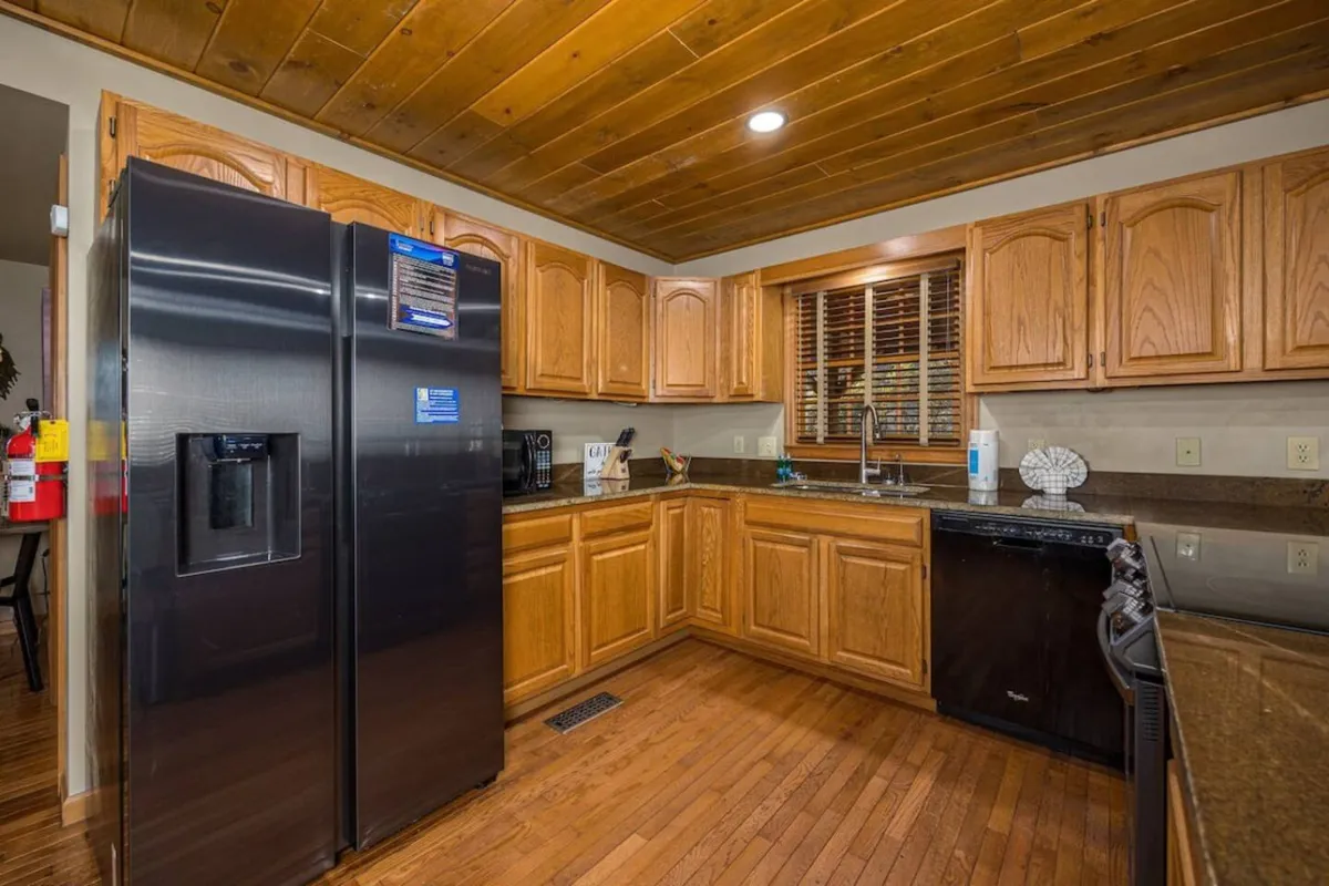 This photo provides a close-up perspective of the kitchen, focusing on a stainless steel refrigerator, wooden cabinetry, granite countertops, and a window that brings in natural light. The bar area with stools is visible, creating a sense of an open, communal cooking and gathering space.