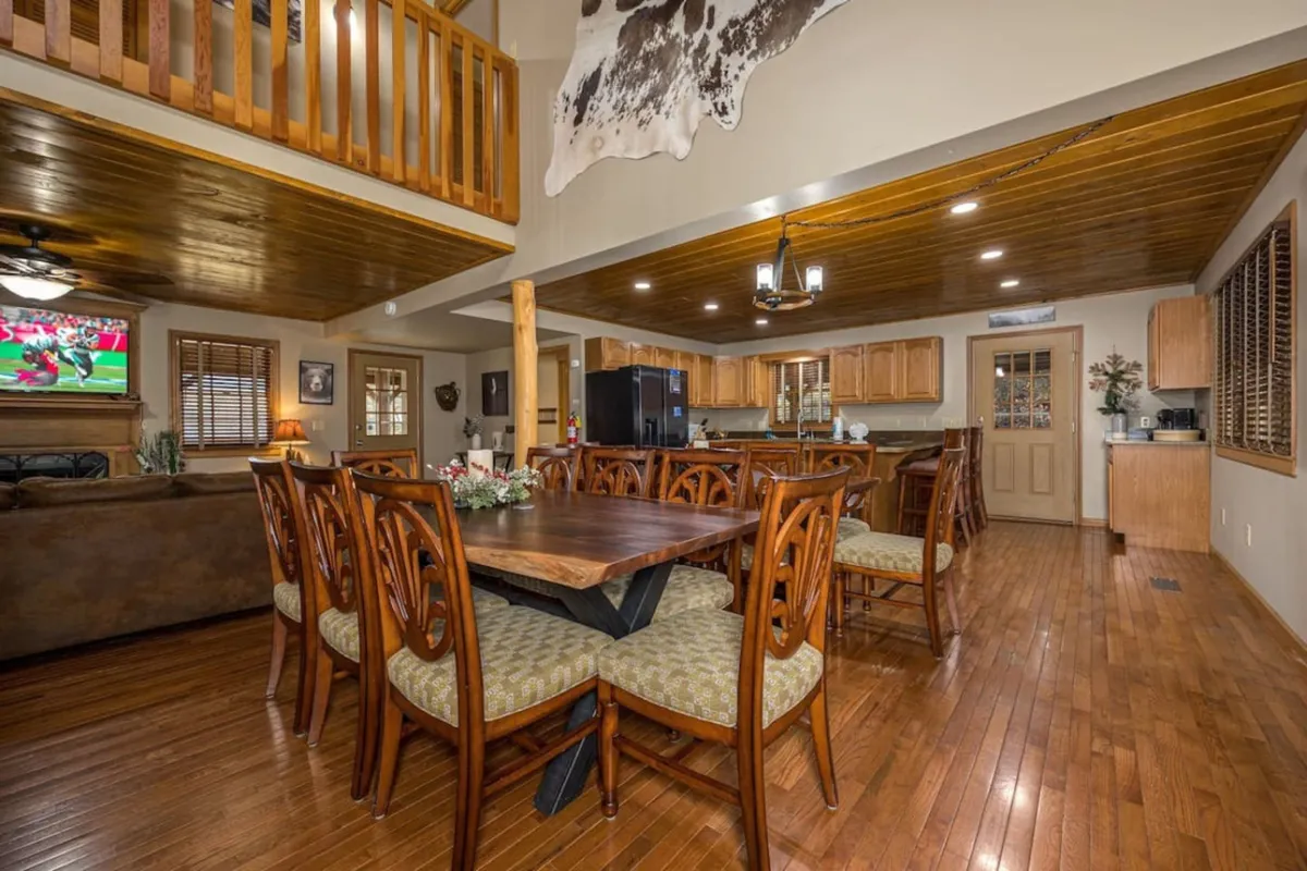 A spacious and inviting open-concept area featuring a large dining table, kitchen, and cozy living room seating. The room is framed by a wooden ceiling and filled with natural light, creating a welcoming atmosphere for large gatherings. Upscale finishes and a TV on the wall add a modern yet warm touch.