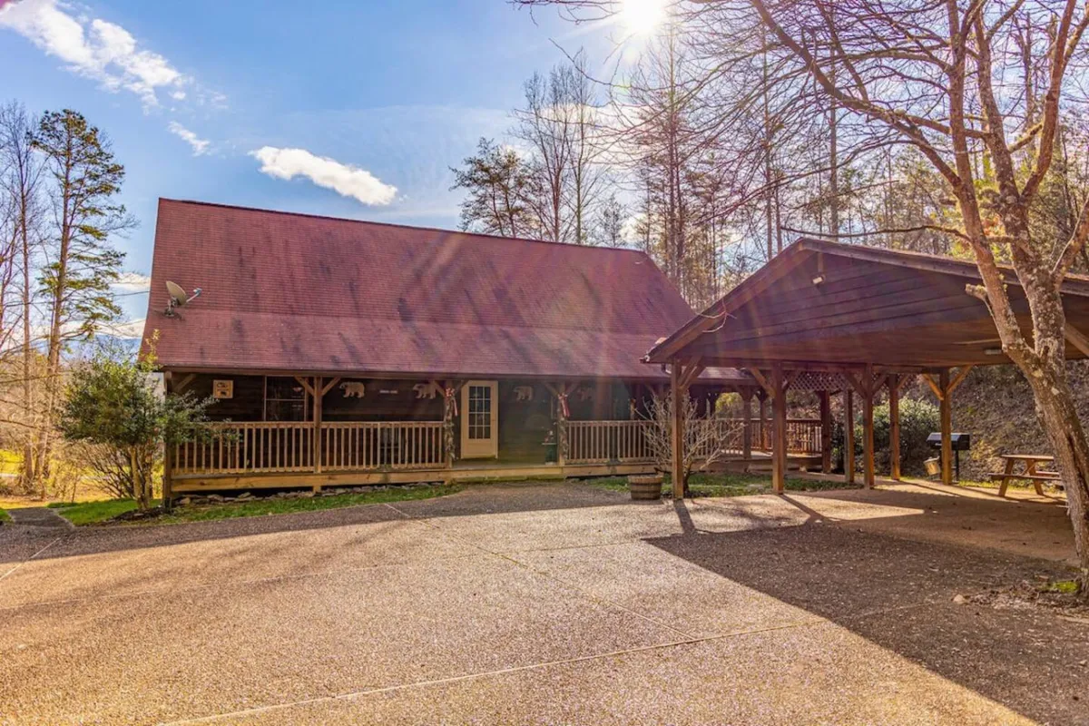 A welcoming log cabin with a distinctive red roof and expansive wooden porch sits amidst lush trees. The porch and surrounding lawn invite group gatherings, outdoor games, and barbecues, evoking a sense of communal fun and relaxation.